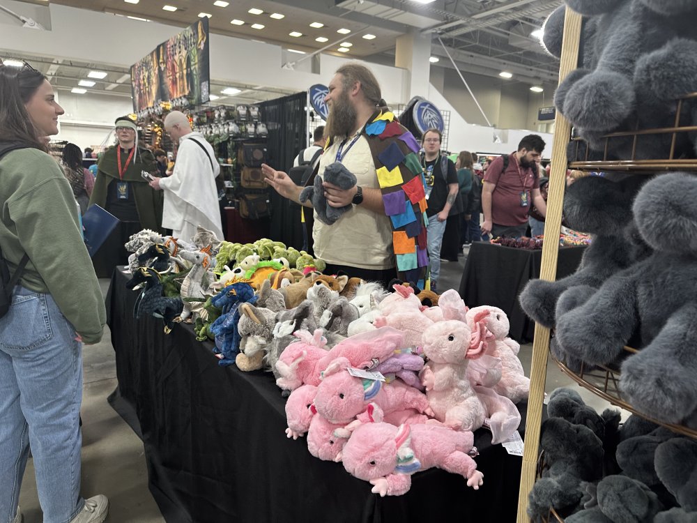 A person in a colorful gleeman's cloak stands over a vendor's table full of plushies