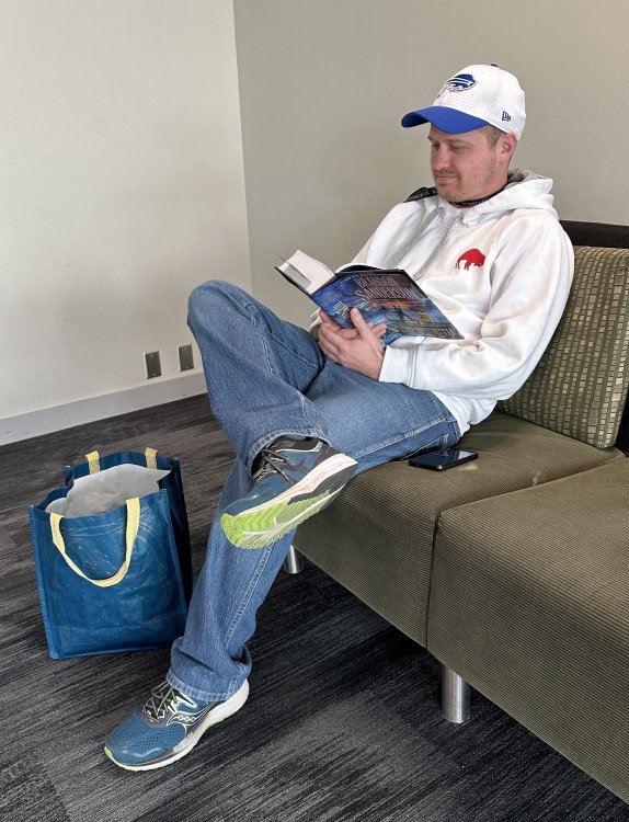 a fan in jeans and a white baseball cap sits on a couch smiling and reading Wind and Truth