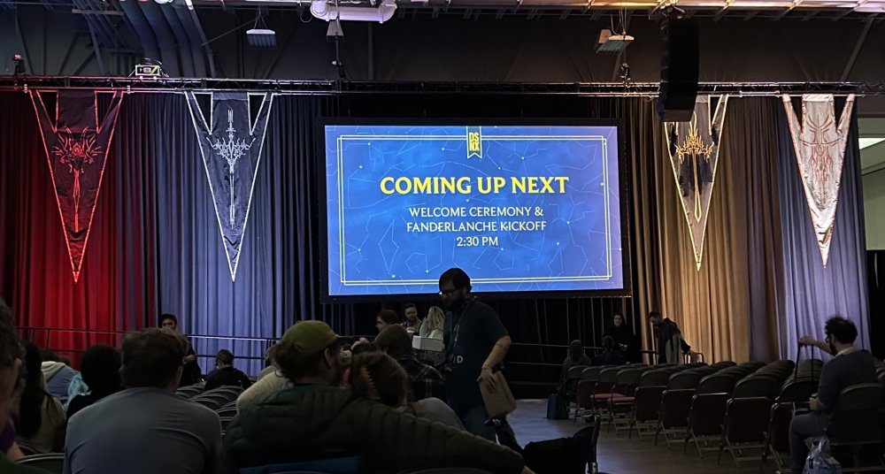 dimly lit auditorium with people in front of a stage with a large tv screen flanked by colorfully lit radiant order banners hanging from the curtain